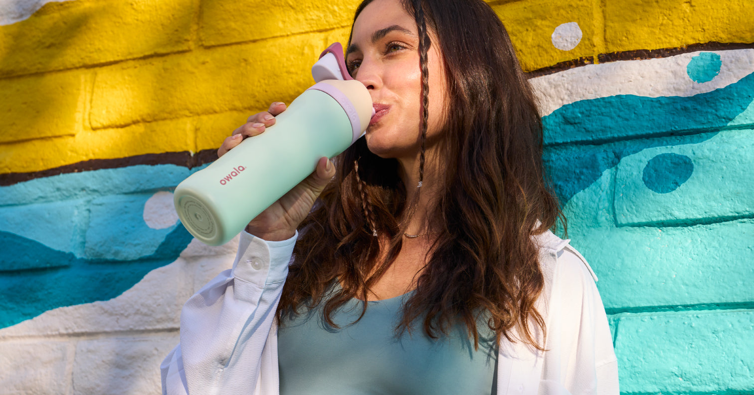 Woman drinks from a mint green Owala bottle against a colorful painted wall
