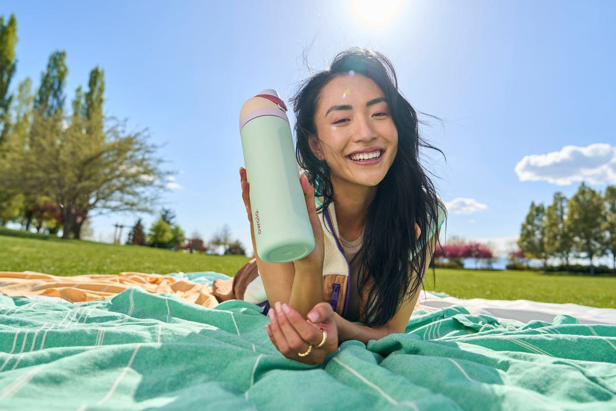 Stylish young woman lying on a teal picnic blanket in a peaceful field during a sunny day, enthusiastically showing off an Owala FreeSip water bottle -- reusable hydration perfect for chill days in the park.