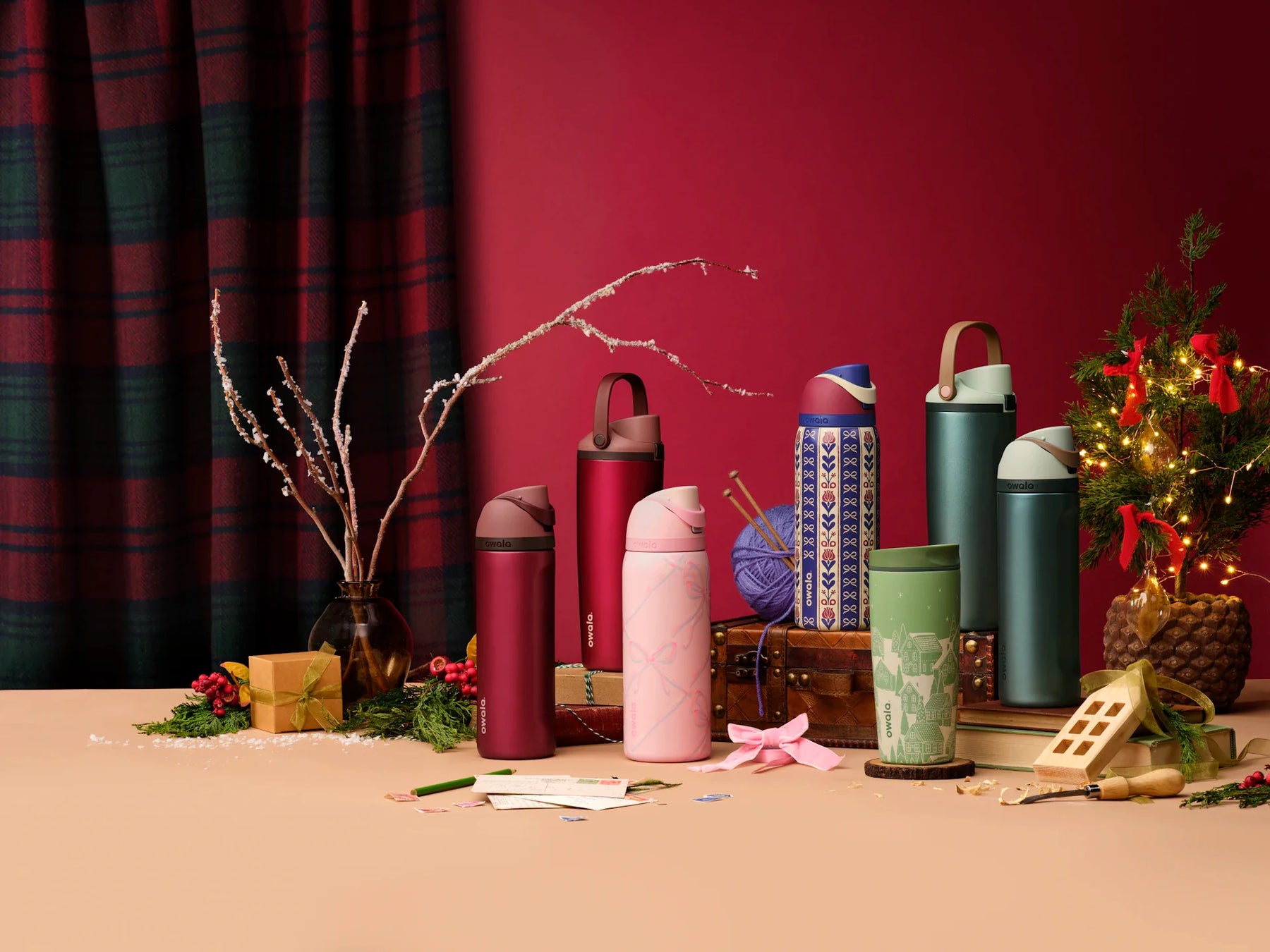 Owala Holiday Collection bottles arranged in front of a red backdrop, with festive elements like ribbon, pine trees, and boxes.