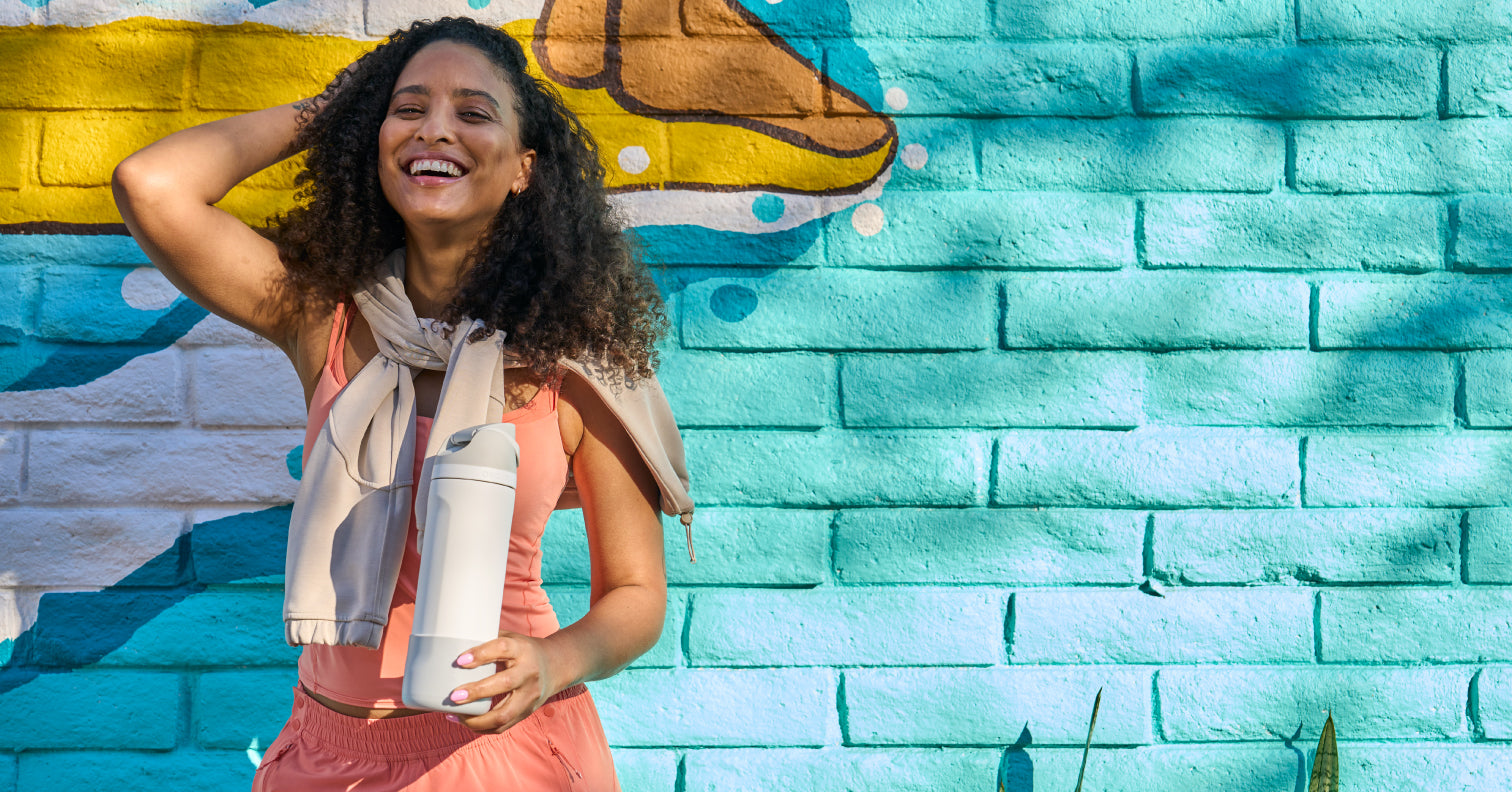 Smiling woman holding a cream-colored Owala bottle in front of a turquoise painted brick wall