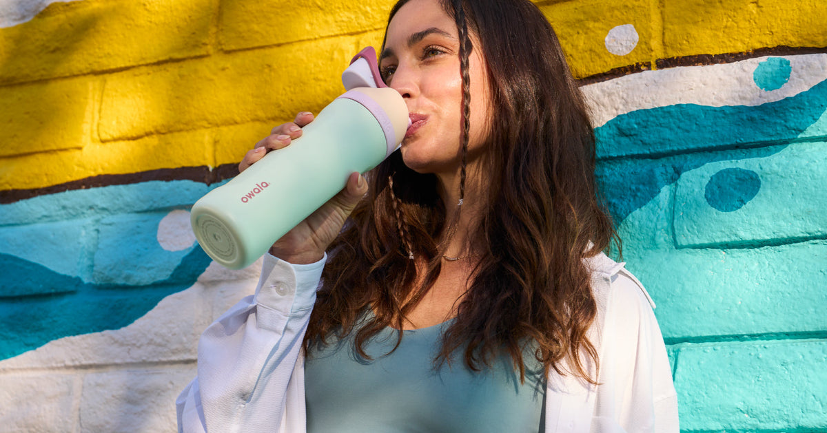 Woman drinks from a mint green Owala bottle against a colorful painted wall