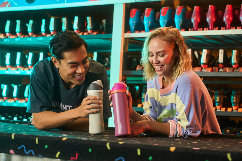 Two friends showing their grey and pink 24oz FreeSip water bottles to each other.