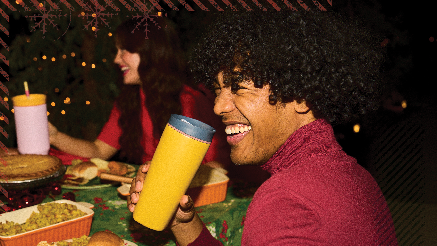Smiling man drinks from a yellow Owala travel mug at a holiday dinner with twinkling lights behind