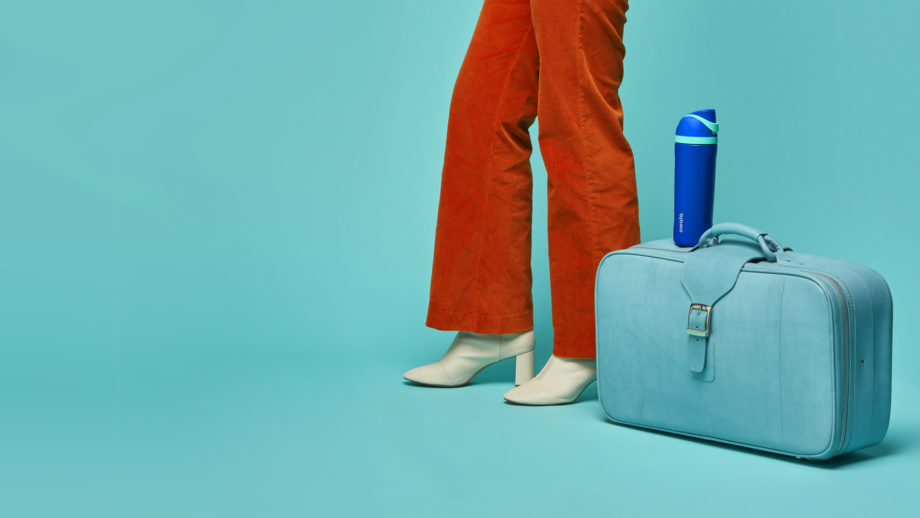 Low-angle shot of a fashionable person wearing wide leg orange jeans, standing next to a teal suitcase with an Owala FreeSip water bottle on top -- gearing up for a road trip!