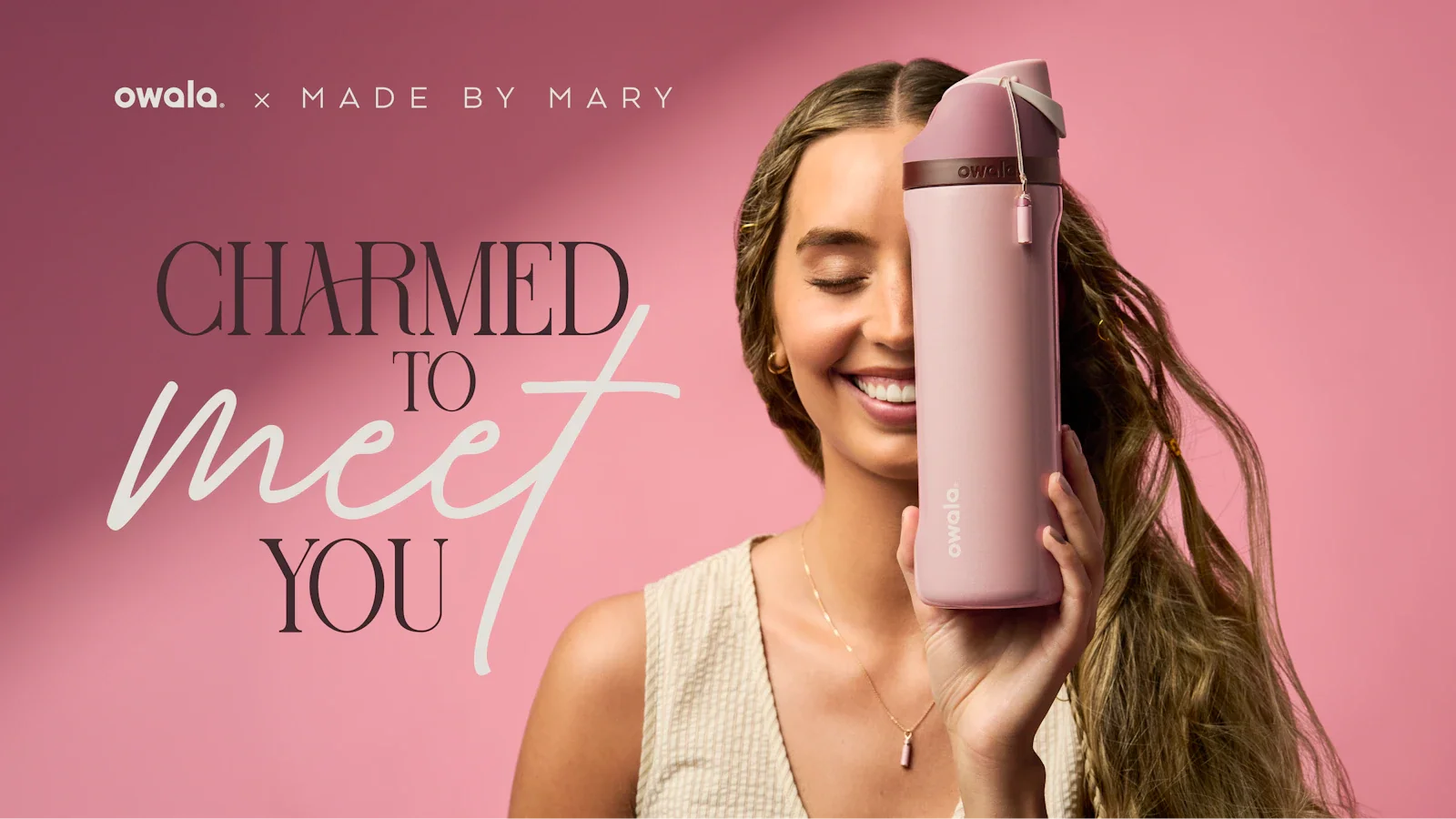 A smiling woman holds a pink water bottle in front of one eye. She wears a matching gold necklace with a pink charm, standing in front of a pink background with the words “Charmed to Meet You” displayed.