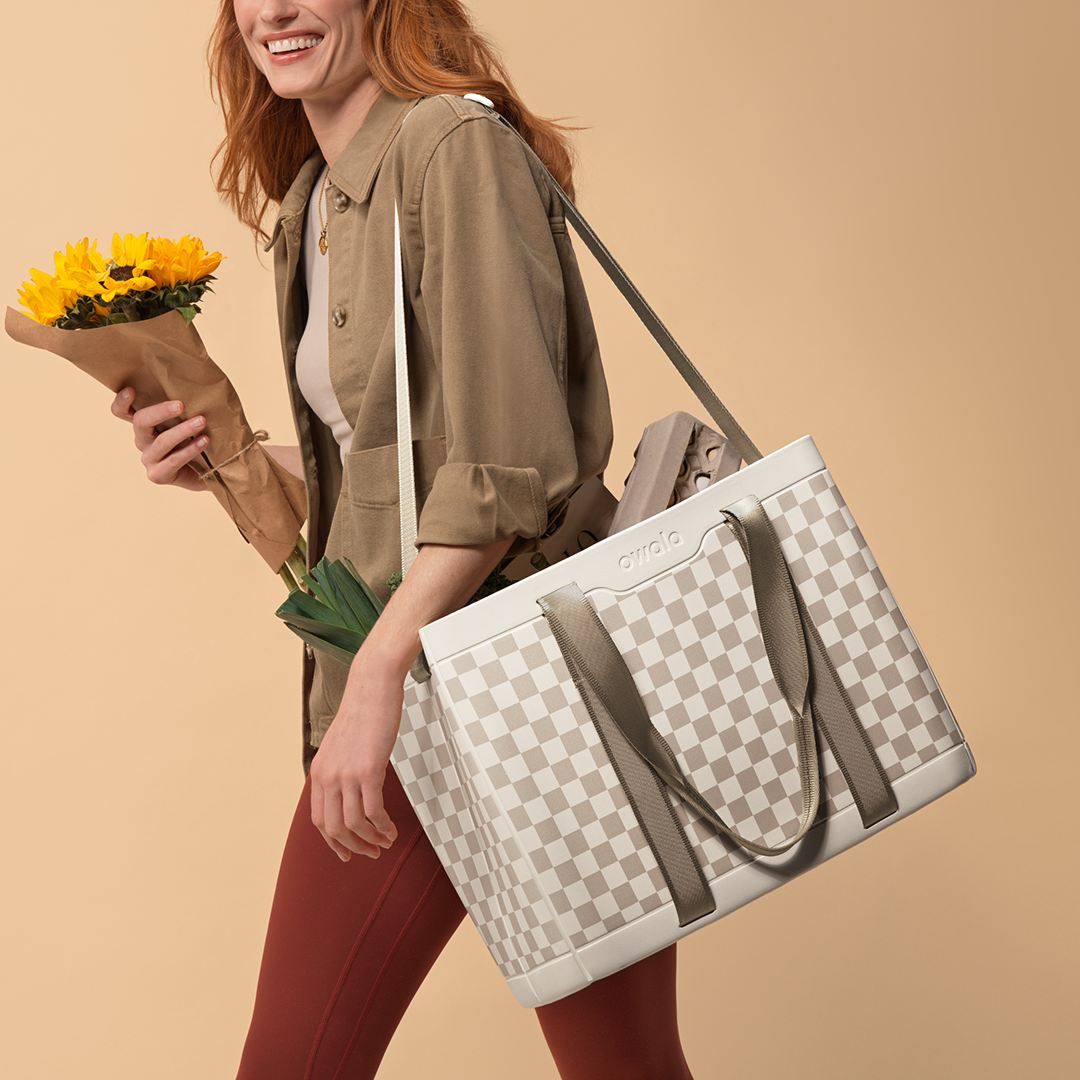 Model holding a sunflower bouquet and carrying the Sun-Day Collapsible Tote in Checkin' In on her shoulder, cream and tan checker body with olive straps